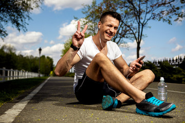 Smiling guy is using headphones and smartphone while sitting on asphalt among green trees. He is having bottle of water besides and demonstrating victory sign. Man is wearing smartwatch and winking