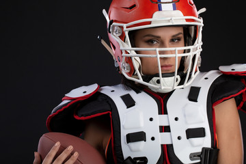 Sportive serious woman in helmet of rugby player holding ball in stuio on black background.