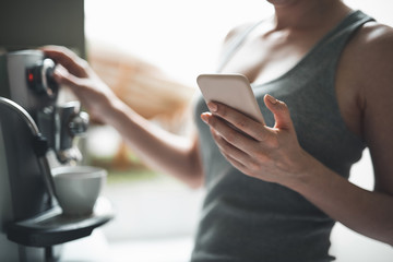Focus on close up female hand holding mobile. Woman is standing by coffee machine and using smartphone to communicate at home