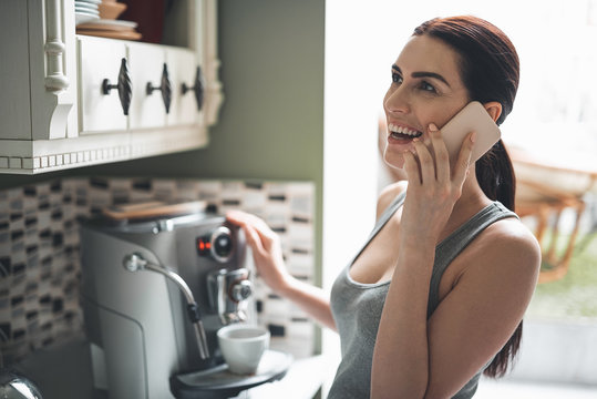 Nice to hear you. Smiling beautiful woman standing by coffee machine and preparing hot drink. She is holding mobile and talking with joy while waiting
