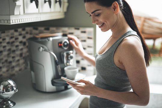 Side view of smiling attractive woman standing by coffee machine with smartphone in hands. She is chatting online with delight