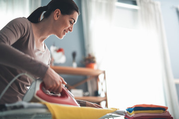 Beautiful housewife concentrated on ironing casual clothes for family. She is smiling and looking at board with content