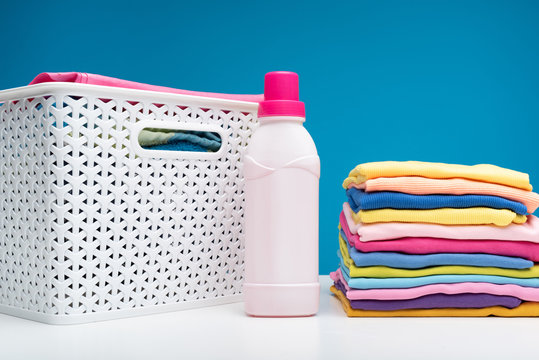 Close up of colorful garments stacked in pile. Bottle of softener and wicker clothesbasket are standing aside
