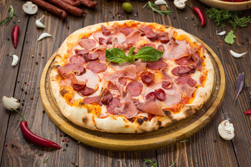 Delicious italian pizza with ham, bavarian sausages and prosciutto decorated by basil leaves on wooden background