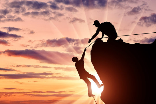 Silhouette of a climber who helps to climb the top of a man, throws him a rope and holds out his hand.
