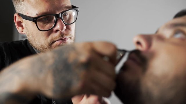 Tattoed barber shears the customer's mustage by using trimmer and comb at the barber shop, man's haircut and shaving at the hairdresser, shaving salon