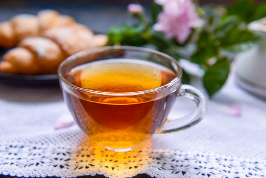 Close up glass cup of hot tea on the vintage napkin with croissants and fresh tea rose flowers on the background. Cozy breakfast, tea time. Selective focus, copy space.