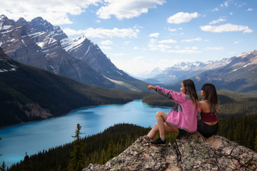 Adventurous female friends sitting on the edge of a cliff overlooking the beautiful Canadian Rockies and Peyto Lake. Taken in Banff National Park, Alberta, Canada. - obrazy, fototapety, plakaty