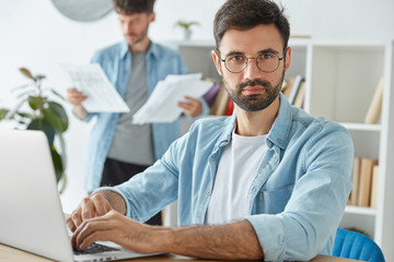 Two young businessmen spend productive morning in office, develop company`s strategy, work with laptop computer and business papers. Bearded young male manager in spectacles works in cabinet