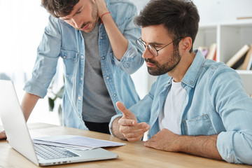 Obraz premium Photo of two young male entrepreneurs work together on financial report, look attentively at documents, creat graphics on laptop computer, dressed in denim fashionble clothes. Business concept
