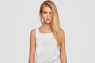Obraz premium Waist up portrait of sexual young female with blonde long hair combed on one side, wears oversized casual white t shirt, shows perfect figure, has healthy pure skin, goes in for sport regularly
