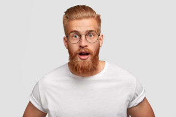 Emotional hipster with stupefied expression, wonders latest news, has thick red beard and mustache, stares at camera, can`t believe his eyes, wears casual white t shirt and round spectacles.