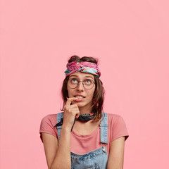 Pretty stylish female hippy in casual wear, has doubtful expression, focused upwards, holds fore finger in mouth, tries to find best solution, looks with indecisiveness, isolated on pink background - obrazy, fototapety, plakaty