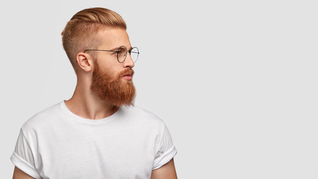 Profile shot of handsome male with trendy hairdo and beard, looks aside with serious expression, has thick red beard, wears round glasses, focused into distance, isolated over white background