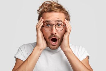 Headshot of handsome unshaven young male looks with terrific expression, keeps hands on head, sees...