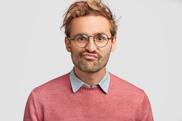 Puzzled displeased male curves lips, looks doubtfully into camera, feels hesitation, dressed in pink sweater, has curly hair, poses against white background. Discontent Caucasian man indoor.