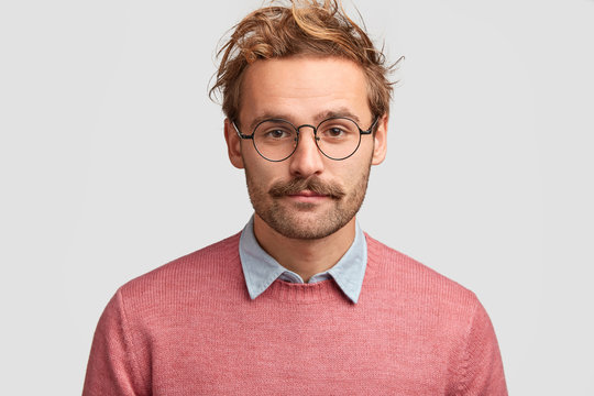 Serious male teacher with confident clever look, has beard and mustache, listens pupil`s answer, wears pink sweater, round glasses, isolated over white background. People, facial expressions concept