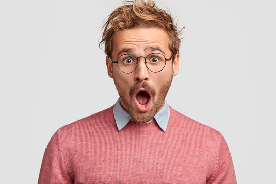 Stressed hipster male has shocked expression, realizes that his car is stolen, keeps mouth widely opened, stares through round spectacles, isolated over white background. People and emotions concept