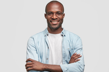 Horizontal shot of prosperous businessman keeps hands crossed, has satisfied expression, looks joyfully at camera, glad to achieve success, dressed in jean jacket, isolated over white background