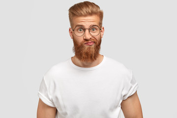 Handsome bearded male purses lips with hesitation, being surprised and puzzled to recieve...