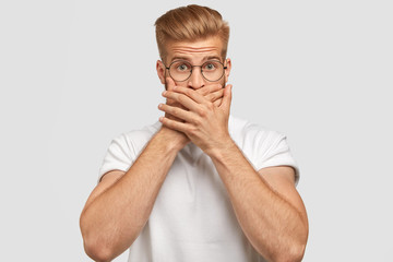 Indoor shot of attractive young male with trendy haircut, covers mouth, being mute, has surprised...
