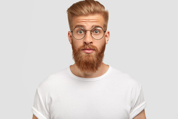 Obraz premium Photo of surprised young male with trendy haircut, has ginger beard and mustache, realizes that girlfriend didn`t come on date, stares with stupefied expression, isolated over white background