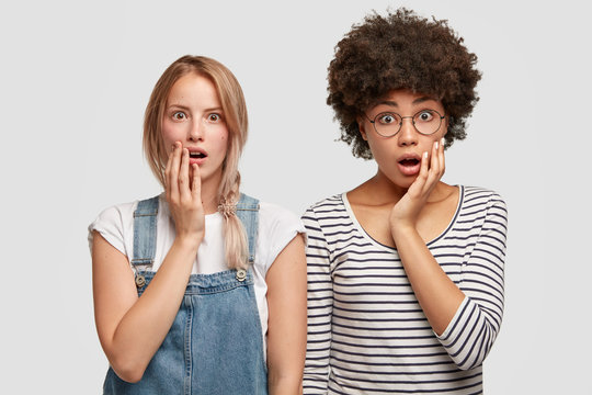 Photo of two suprirsed women stares with widely opened mouthes at camera, dressed in fashionable clothes, can`t believe in something awful, pose against white background. Multiethnic females