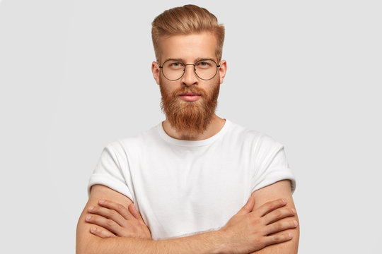 Studio shot of serious self assured male architect keeps hands crossed, has ginger thick beard and mustache, listens attentively client, stands against white background. Confident stylish hipster