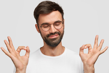 Pleased attractive man with thick dark beard, makes okay gesture with both hands, shows his approval, closes eyes, dressed casually, isolated on white background. Body language and lifestyle concept