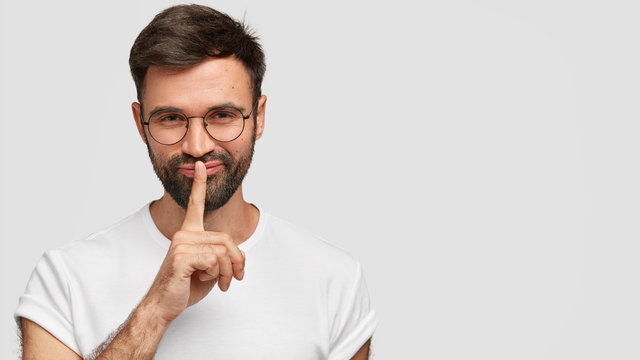 Cheerful mysterious bearded male with dark hair and stubble, makes silence gesture, tells privare information, shares good news with friend, dressed in white t shirt. Monochrome. Secrecy concept