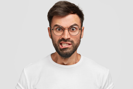 Displeased bearded male frowns face with displeasure, has irritated expression, raises eyebrows and clenches teeth, being discontent, argues with enemy, wears spectacles. Negative facial expressions