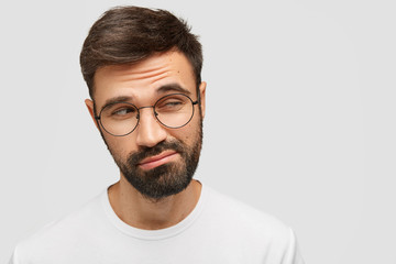 Clueless doubtful male with dark thick beard, looks with hesitation aside, raises eyebrows in bewilderment, has pensive expression, isolated over white background with copy space for advertisement - obrazy, fototapety, plakaty