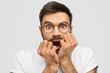 Human emotions and feelings concept. Scared unshaven male bites nervously fists, opens eyes widely, feels anxious, being scared of something terrified, wears round spectacles and white t shirt