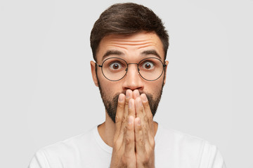Surprised young attractive Caucasian male covers mouth and looks with unbelievable expression,...