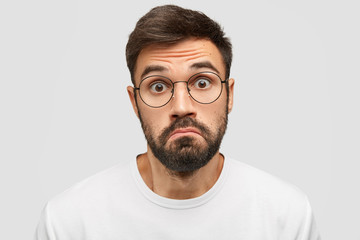 Obraz premium Doubtful bearded male purses lips with hesitation, stares at camera with scared expression, feels cluless while recieves unexpected offer from friend, isolated over white background. Facial expression