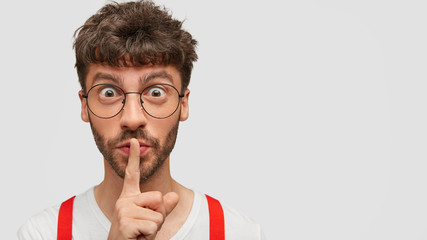 Shush be silent! Mysterious bearded guy with surprised expression, keeps fore finger on lips as...