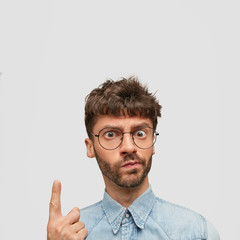 Obraz premium Indignant bearded male looks angrily at camera, has discontent expression, points upwards with index finger, shows blank space for your advertisement, wears denim elegant shirt and big glasses