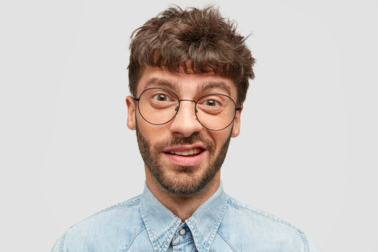 Photo of funny male with stubble, has indecisive and curious look, frowns face, looks directly at camera, dressed in denim shirt, isolated over white background. People and facial expressions concept