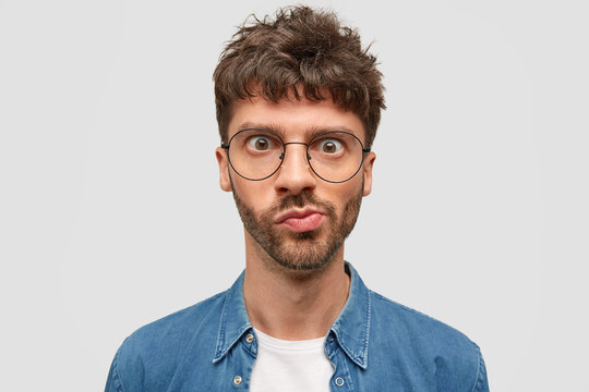 Headshot of puzzled bearded young man looks in wonderment, curves lips, has widely opened eyes, dark wavy hair, recieves unexpected news, thinks over something, stands against white studio wall