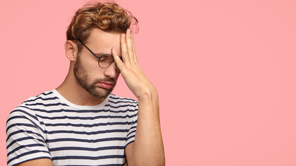 Attractive tired overworked male entrepreneur sighs with tiredness, keeps hand on forehead, suffers from headache, hangs head, stands in profile against pink background. Omg, I am fed up of everything