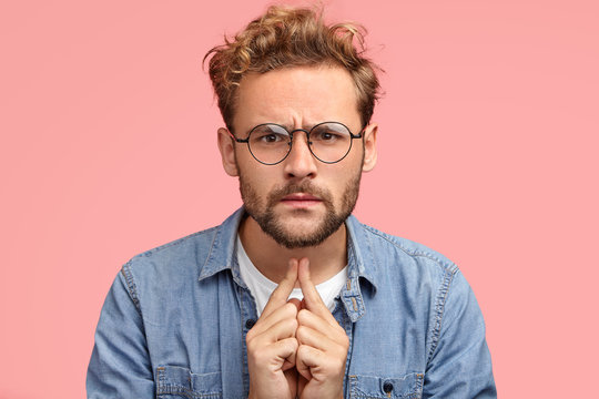 Headshot of serious intelligent young male proffesor listens something with attentive look, has scrupulous gaze directly into camera, dressed in fashionable outfit, isolated over pink background