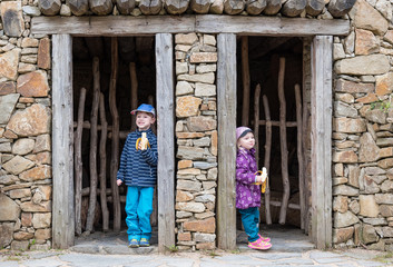 Fototapeta na wymiar Two children are standing in different doors of an old stone house having banana for a snack