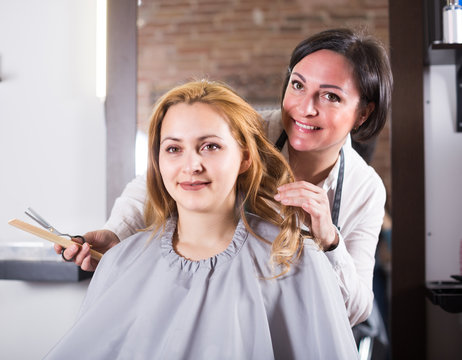 Mature hairdresser is doing fashionable haircut to young woman with scissors and comb