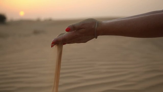 An amazing view of nice woman`s hands with red makeup keeping a handful of desert sand and permitting it to fall in Dubai at a shimmering sunset 