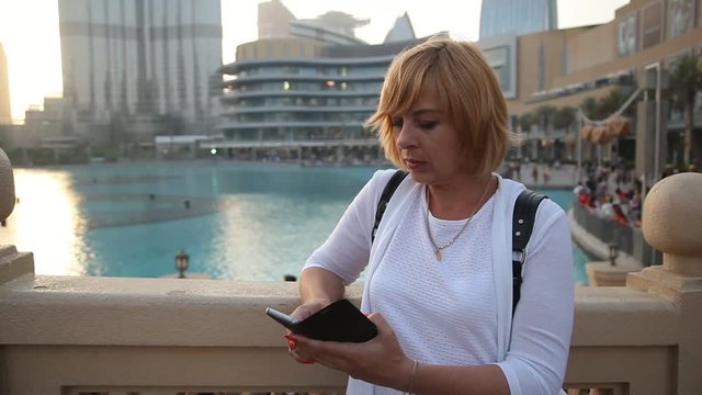 Portrait of a modern female backpacker in Dubai. She stands on a water channel quay, surfs her smartphone leaning on fence in summer