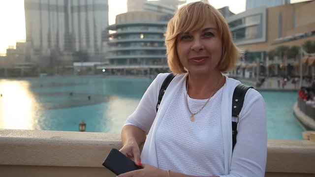 Portrait of a cheery blond woman in white blouse standing on an embankment and smiling in Dubai in summer. Burj Khalifa is behind her back