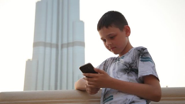 An exciting view of a smart boy surfing the net on his mobile and smiling cheerfullyon a water channel quay. Burj Khalifa is seen too.