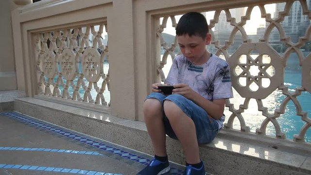 An emotional view of a 10-year-old boy plunging in the virtual reality of his smartphone while sitting on a water channel quay in Dubai in summer