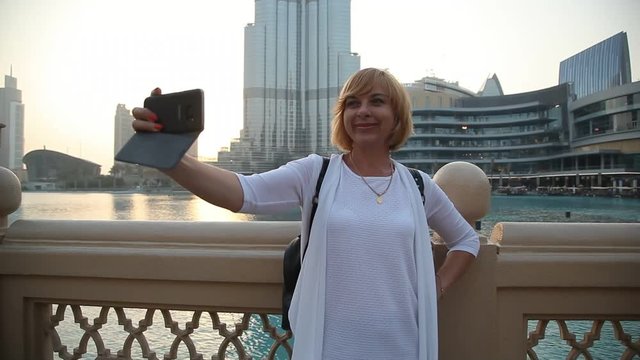 Portrait of a happy blond woman standing at a water channel and taking a selfie with Burj Khalifa, the highest building in the world, in summer.
