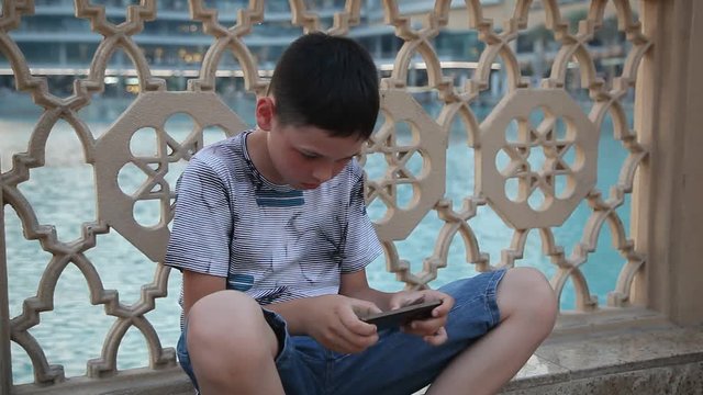 An original view of a 10 year old boy playing video games at his smartphon. He sits at a wooden fence being in a T-shirt and shorts in summer
